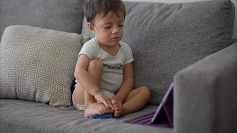 Süßes-Junges-Lateinisches-Kleinkind-In-Einem-Hellblauen-Babyspielanzug,-Der-Auf-Einer-Grauen-Couch-Sitzt-Und-Cartoons-Auf-Einem-Lila-Tablet-Anschaut