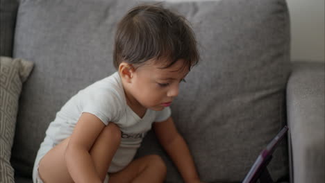 Beautiful-latin-baby-boy-in-a-light-blue-baby-romper-sitting-on-a-grey-couch-watching-a-show-on-a-purple-tablet