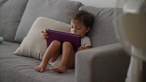 Cute-little-latin-baby-boy-in-a-light-blue-baby-romper-sitting-on-a-grey-couch-watching-a-movie-on-a-purple-tablet