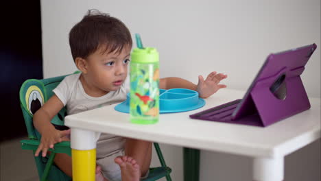 Cute-latin-baby-boy-enjoying-a-movie-on-a-purple-tablet-while-having-breakfast-sitting-on-a-white-children-table