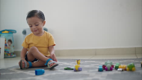 Young-cute-latin-toddler-sitting-on-the-floor-on-a-grey-carpet-making-faces-and-playing-with-his-car-toys