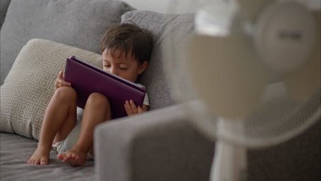Cute-little-latin-toddler-in-a-light-blue-baby-romper-sitting-on-a-grey-couch-enjoying-a-movie-on-a-purple-tablet