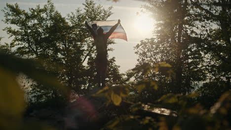 Frog's-eye-perspectice-on-man-holding-Slovenian-flag-towards-the-morning-sun-on-peak-of-mountain-Slivnica,-between-the-trees