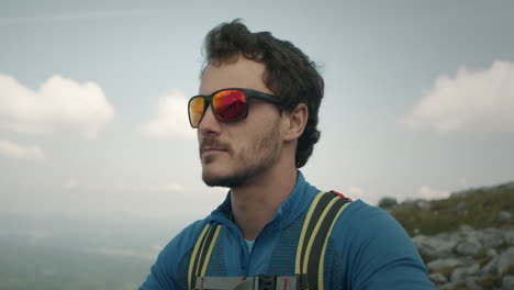 Close-shot-of-a-hiker-with-a-green-backpack-and-dark-sunglasses-with-red-lenses-looking-at-the-beautiful-view-from-peak-of-the-mountain