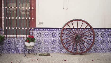 typical-Spanish-yard-with-an-old-wheel,-a-few-plants-with-red-flowers-and-blue-tiles