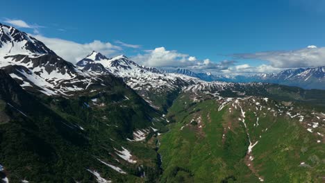 Fliegen-über-Dem-Kenai-Fjords-National-Park-Mit-üppigen-Grünen-Bergen,-Die-Teilweise-Von-Schnee-Bedeckt-Sind,-Unter-Blauem-Bewölktem-Himmel-In-Alaska,-Usa