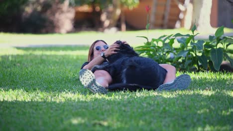 A-young-caucasian-woman-lying-on-the-grass-plays-with-her-black-dog-in-the-garden-of-a-house-on-a-sunny-day