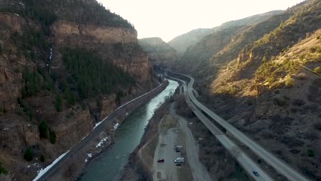 aerial-drone-flying-backwards-in-a-canyon-with-a-highway,-river-and-flying-higher-up-as-the-shot-goes-on