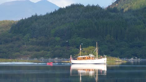 Wooden-Boat-with-small-Zodiac-boat-sailing-on-a-Lake-in-Scotland-with-Forest-and-Mountains-in-background