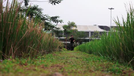 Asian-man-wearing-black-shirt-and-long-camo-cargo-pants,-running-away-from-camera-in-a-paddy-field,-in-low-angle-shot
