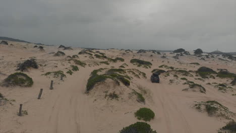 Aerial-following-4x4-vehicle-driving-around-sandy-beach---grassy-dunes