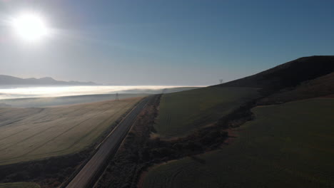 Aerial-View-of-Misty-Plains-and-an-Empty-Road-in-the-Morning-Hours-with-Mountains-in-the-Background