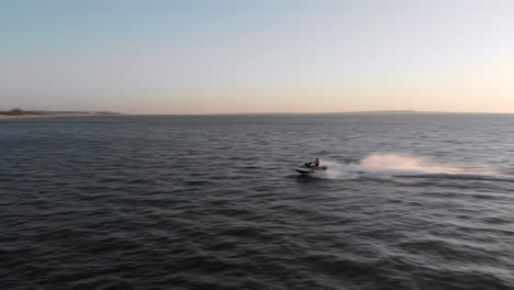 A-Woman-Rides-a-Jet-Ski-at-High-Speed-on-an-Open-Ocean-at-Sunset,-Drone-Tracking-Shot