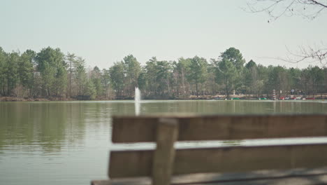 Focus-pull-to-the-left-side-of-a-wooden-bench-in-front-of-a-lake-with-a-water-fountain-in-the-middle