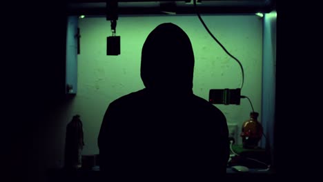 Silhouette-of-a-mysterious-hacker-wearing-a-hoodie-playing-his-computer-and-his-phone-full-of-green-programming-codes-in-a-dark-room