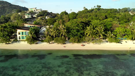 Sideways-flight-of-a-drone-looking-at-a-beach-in-Thailand-with-houses-and-a-hotel-on-the-background