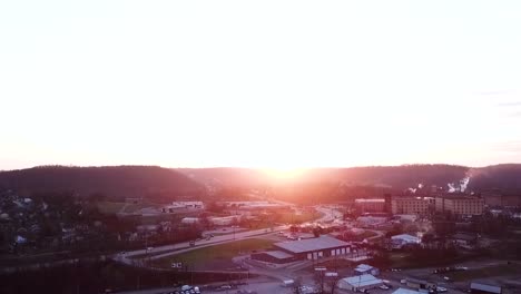 An-aerial-zoom-pan-of-a-beautiful-golden-sun-setting-over-a-small-city-area-with-a-smoke-producing-bourbon-distillery-in-the-background
