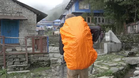 Joven-En-Cámara-Lenta-Caminando-Por-El-Pueblo-De-Nepal-Con-Mochila-Naranja-Cubierta-De-Lluvia,-Annapurna
