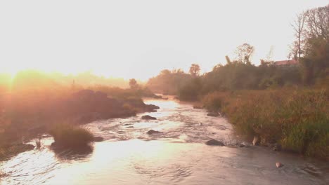 Drone-video-with-moving-forward-along-a-river-with-morning-sun-rays-glittering-off-the-water-on-a-foggy-morning