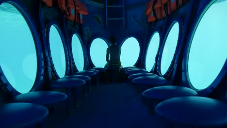 Push-in-on-a-boy-sitting-all-alone-facing-away-as-he-looks-out-the-porthole-of-a-submarine-under-a-dreamlike-blue-glow