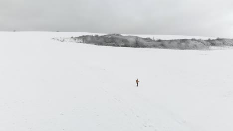 Aerial-Push-out-shot-of-male-person-walking-down-hill-in-wide-snowy-winter-landscape