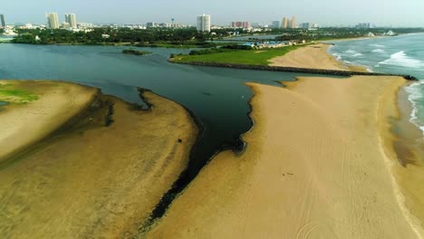 Drone-footage-of-Beach-with-a-backwater-opening-to-it-with-a-city-skyline-with-high-rises-in-backdrop