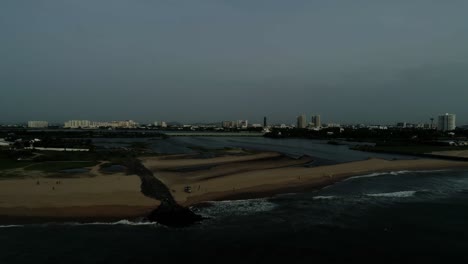Drohnenaufnahmen-Eines-Strandes-Mit-Einem-Sich-öffnenden-Stauwasser-Und-Einer-Skyline-Der-Stadt-Mit-Hochhäusern-Im-Hintergrund