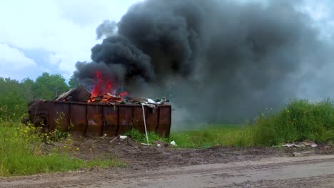 Dicker-Dunkler-Rauch,-Der-Aus-Einem-Brennenden-Müllcontainer-Voller-Müll-Auf-Ländlichem-Ackerland-Aufsteigt