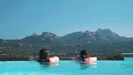 Two-young-girls-relax-in-an-infinity-pool-looking-at-a-picturesque-Sardinian-mountain-scape