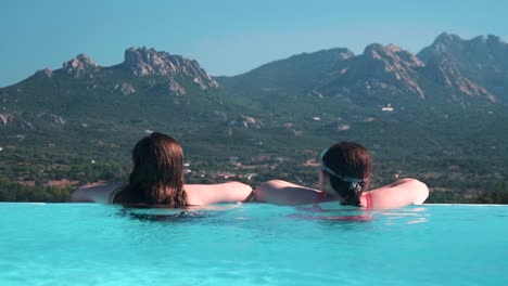 Two-young-girls-relax-in-an-infinity-pool-looking-at-a-picturesque-Sardinian-mountain-scape