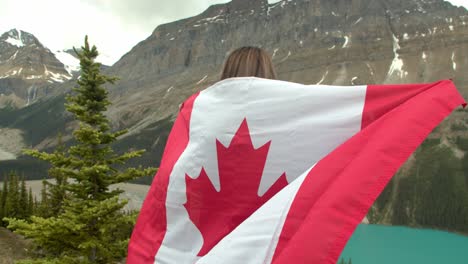 Frau,-Die-Oben-Auf-Einer-Klippe-Steht-Und-Die-Kanadische-Flagge-Vor-Felsigen-Bergen-Und-Einem-Blauen-Gletscherfluss-Hält