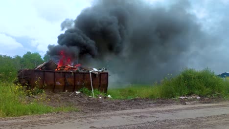 Dicker-Dunkler-Rauch,-Der-Aus-Einem-Brennenden-Müllcontainer-Voller-Müll-Auf-Ländlichem-Ackerland-Aufsteigt
