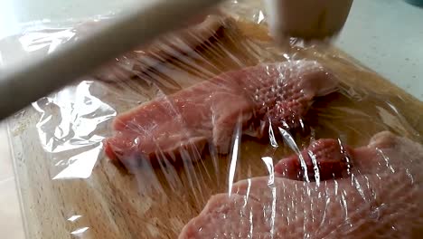 Tenderizing-pork-loin-with-a-wooden-meat-hammer-through-cling-film-on-a-wooden-cutting-board,-SIDE-VIEW-SLOMO