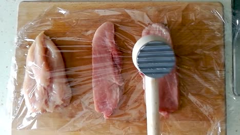 Tenderizing-pork-loin-with-a-wooden-meat-hammer-through-cling-film-on-a-wooden-cutting-board,-OVERHEAD-SLOMO