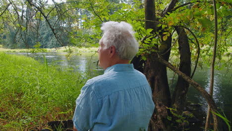 Older-man-sits-by-river,-tall-grass,-trees,-enjoying-nature,-serene