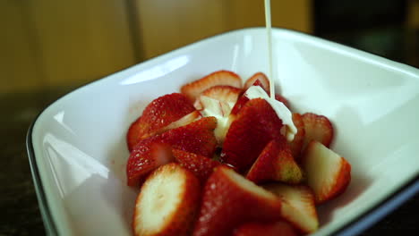 Heavy-whipping-cream-slowly-dripping-over-ripe-strawberries-in-a-white-bowl-on-a-kitchen-counter,-closeup-slow-motion