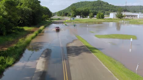 4k-rising-aerial-view-of-a-car-driving-through-flood-water-going-over-a-road-outside-of-a-small-town