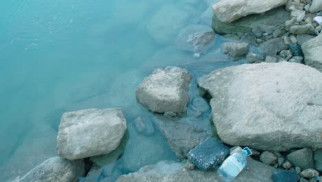 Plastic-bottle-and-garbage-pollution-on-rocks-at-edge-of-water,-Pan