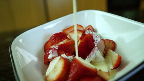 Heavy-whipping-cream-slowly-dripping-over-ripe-strawberries-in-a-white-bowl-on-a-kitchen-counter,-closeup-slow-motion