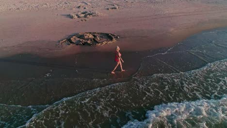 AERIAL:-Circling-Confident-Young-Woman-slowly-walking-on-the-beach-in-beautiful-sunset-light-with-waves-touching-her-feet-[4K