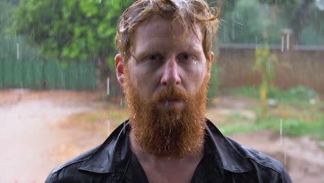 Slow-motion-portrait-shot-of-a-bearded-ginger-man-standing-in-the-pouring-rain-and-getting-soaked-while-looking-into-the-camera