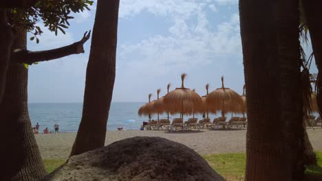 Gimbal-Aufnahme-Des-Tropischen-Strandes-Von-Marbella-Mit-Tikki-Sonnenschirmen-Im-Hintergrund-Und-Felsen-Und-Palmen-Im-Vordergrund