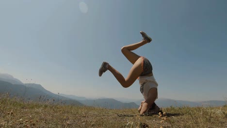 Young-girl-trying-to-make-a-yoga-headstand-on-a-mountain,-on-a-sunny-day