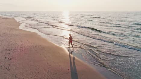 AERIAL:-Circling-Confident-Young-Woman-walking-on-the-beach-in-beautiful-sunset-light-with-waves-touching-her-feet-[4K