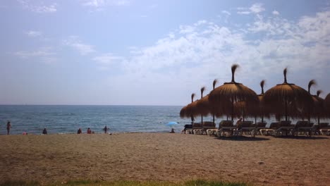 Bewegungszeitraffer-Von-Marbella-Beach-Mit-Tikki-regenschirmen-Und-Wolken,-Die-Sich-Im-Hintergrund-Bewegen,-Sommerferien