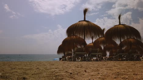 Bewegungszeitraffer-Von-Marbella-Beach-Tiki-Sonnenschirmen-Im-Vordergrund-Und-Wolken,-Die-Sich-An-Einem-Sommertag-Im-Hintergrund-Bewegen