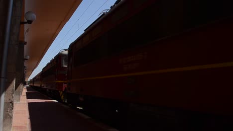 Red-train-engine-arriving-at-Bulgarian-railway-station-platform-on-a-sunny-day-stopping-and-carriage-door-starts-to-open