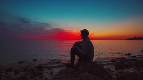 4K-UHD-Cinemagraph-of-a-young-man-with-a-baseball-cap-sitting-by-the-seaside-at-sunset-in-Croatia-with-a-dramatic-red-and-blue-evening-sky