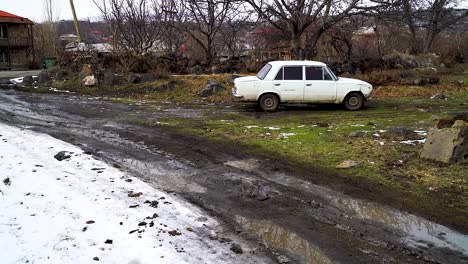 Altes-Weißes-Auto-Im-Sowjetischen-Stil-Biegt-Misstrauisch-Von-Der-Schneebedeckten-Straße-Auf-Einen-Gefrorenen-Schmutzfleck-Ab,-Der-Von-Bäumen-Bedeckt-Ist