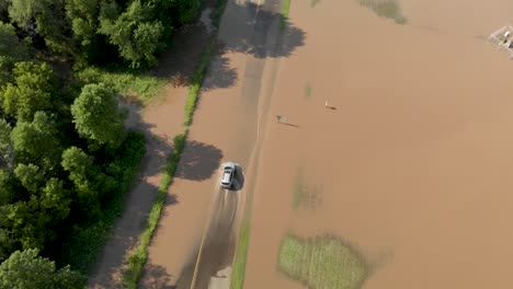 4k-aerial-following-view-of-a-car-driving-through-flooding-on-a-road-outside-of-a-town